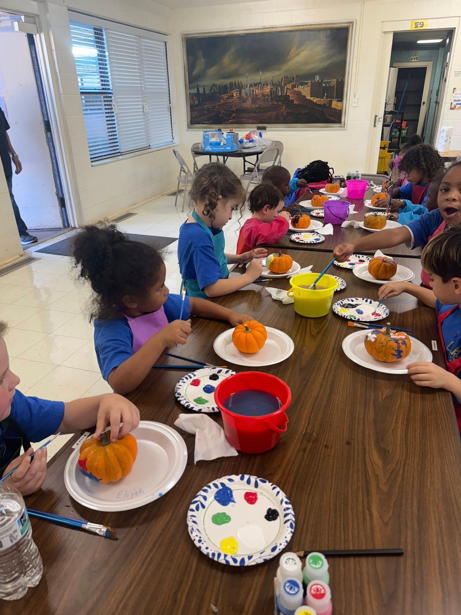 Children seated around a long table, eating snacks and using colorful plates in a bright room.