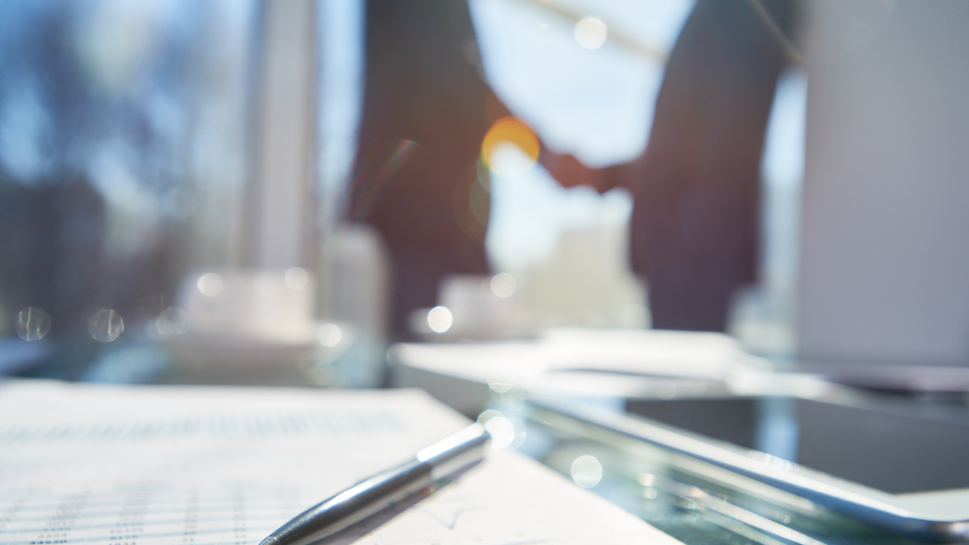 Business people shaking hands in an office, documents and a pen in the foreground.