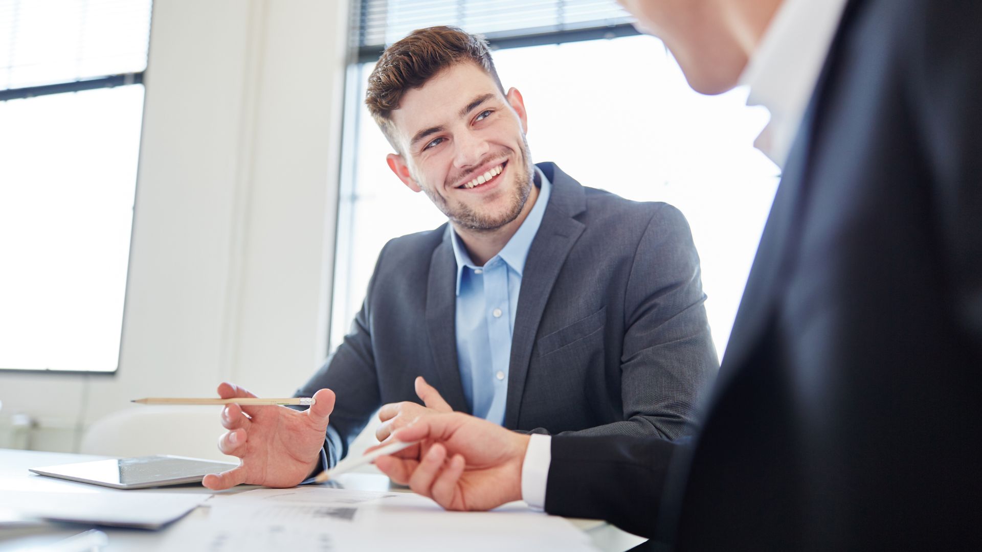 Man in a suit smiling, gesturing with pen during a meeting. Another person in a suit is partially visible.