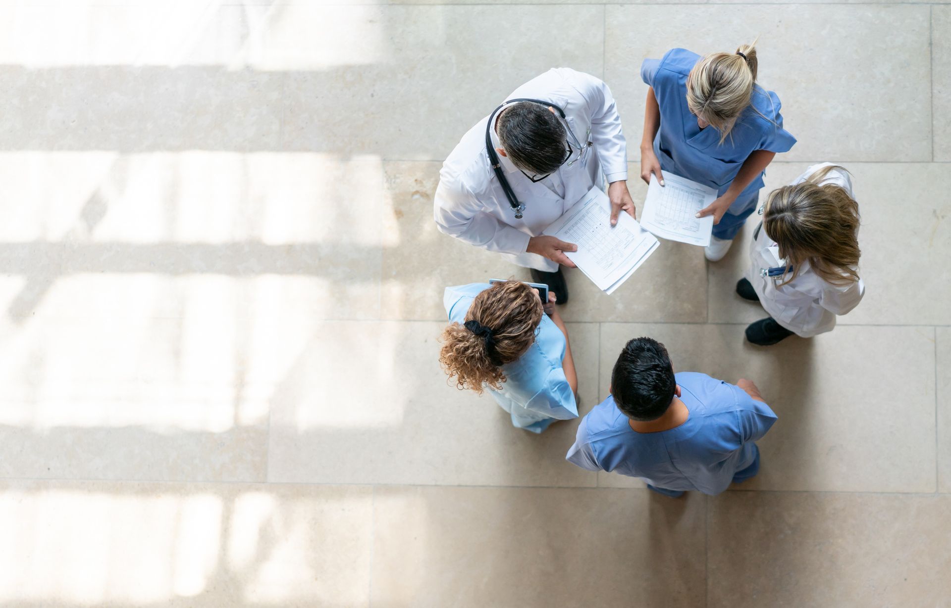 Un team medico riunito discute le cartelle cliniche dei pazienti su un pavimento piastrellato beige.