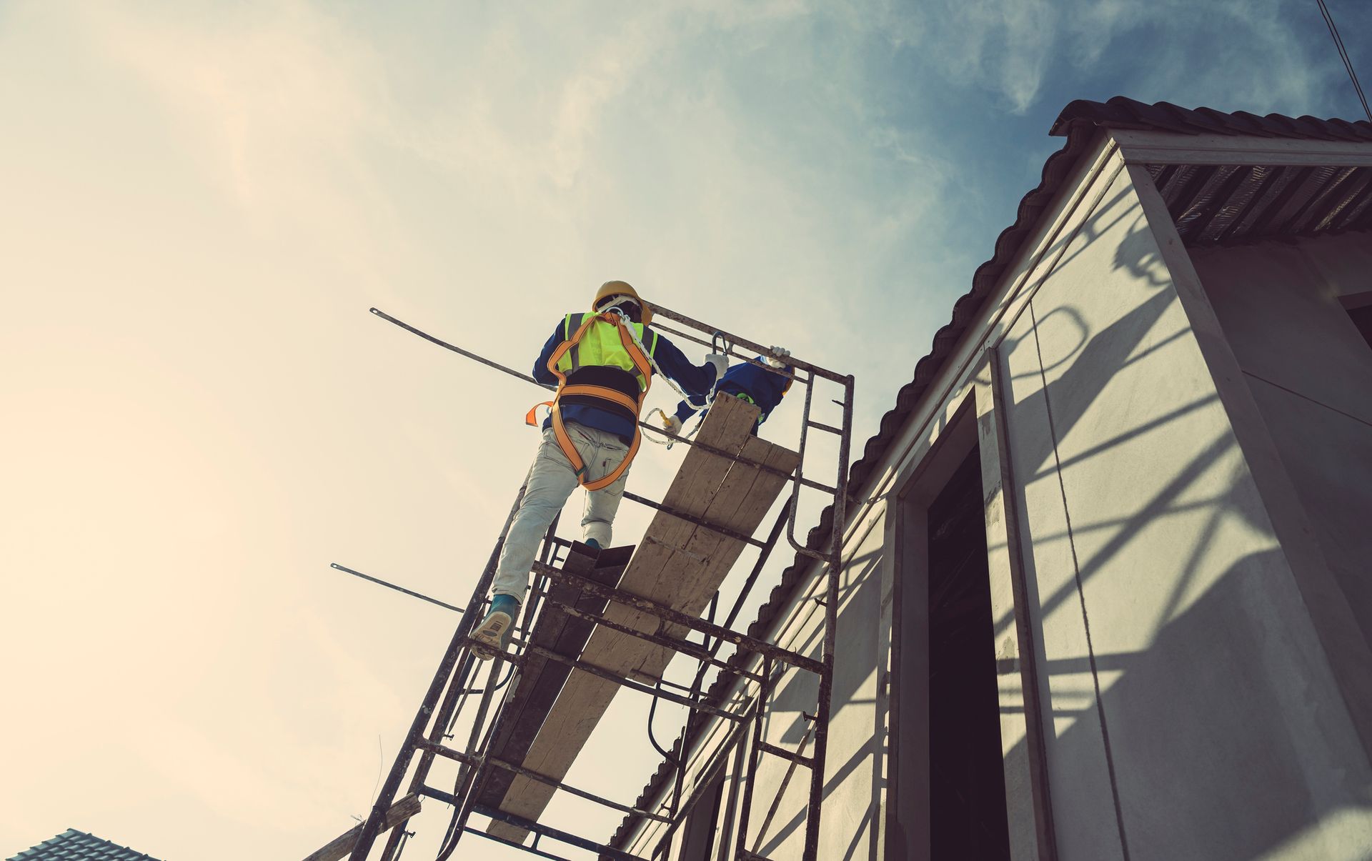Operaio edile su un'impalcatura, con indosso l'equipaggiamento di sicurezza, mentre lavora su un edificio contro un cielo azzurro.