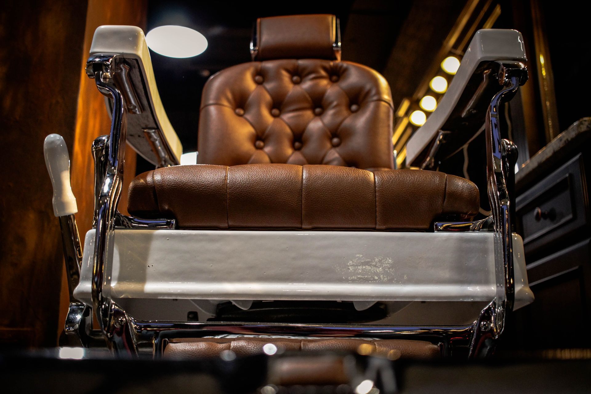 A brown, tufted leather barber chair with chrome armrests and a white base, centered in a dimly lit, classic shop.