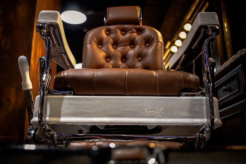 A brown, tufted leather barber chair with chrome armrests and a white base, centered in a dimly lit, classic shop.
