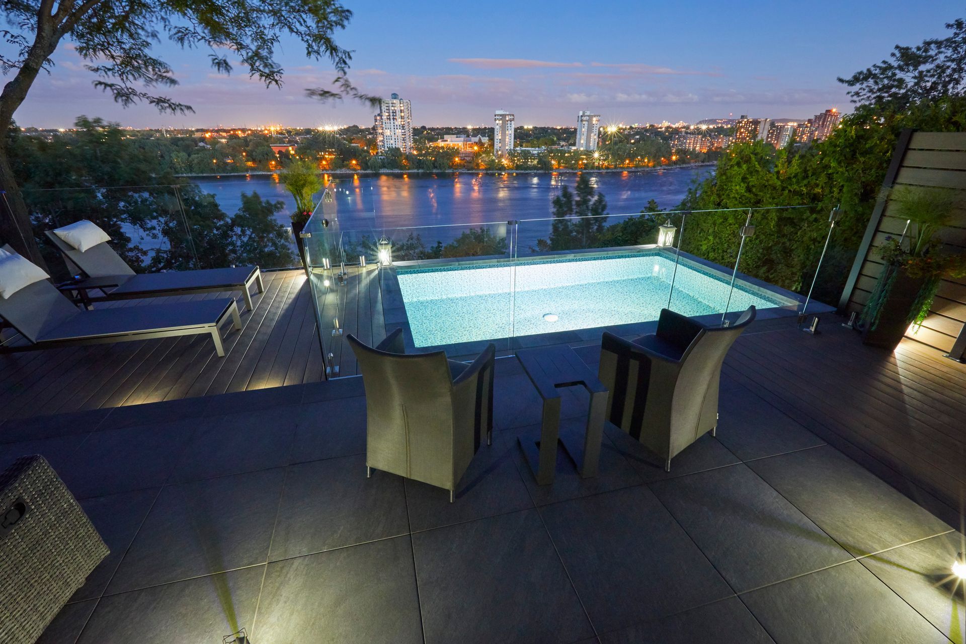 Poolside view of a rectangular pool, chairs, and a lake with a dock, surrounded by green grass and trees.