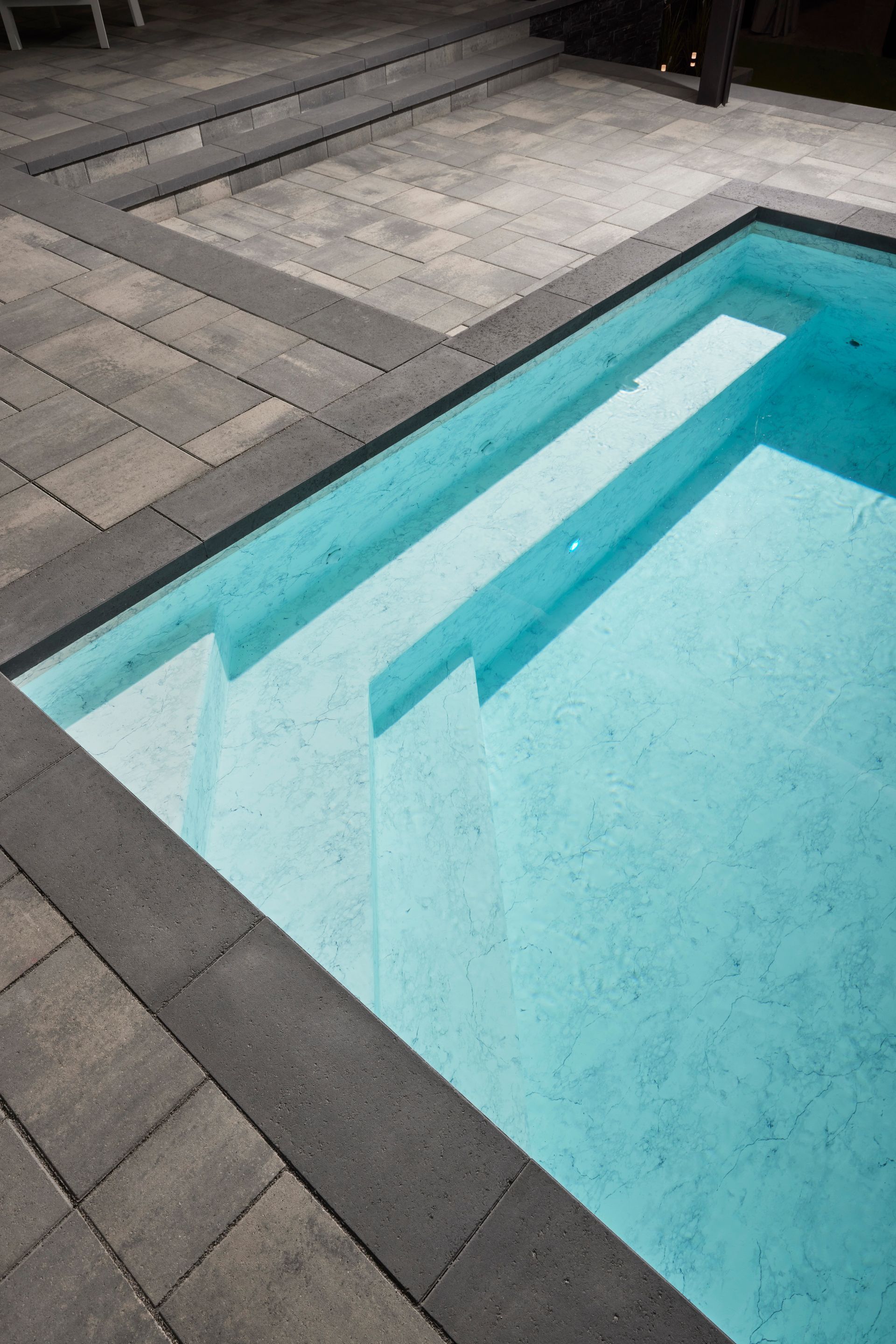 Pool with built-in steps, surrounded by gray paving stones. Turquoise water reflects sunlight.