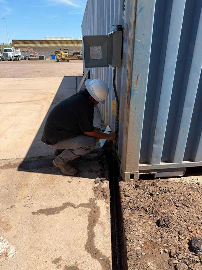 Electrician crouching beside a corrugated metal storage container, using tools near the base of a wall outdoors.