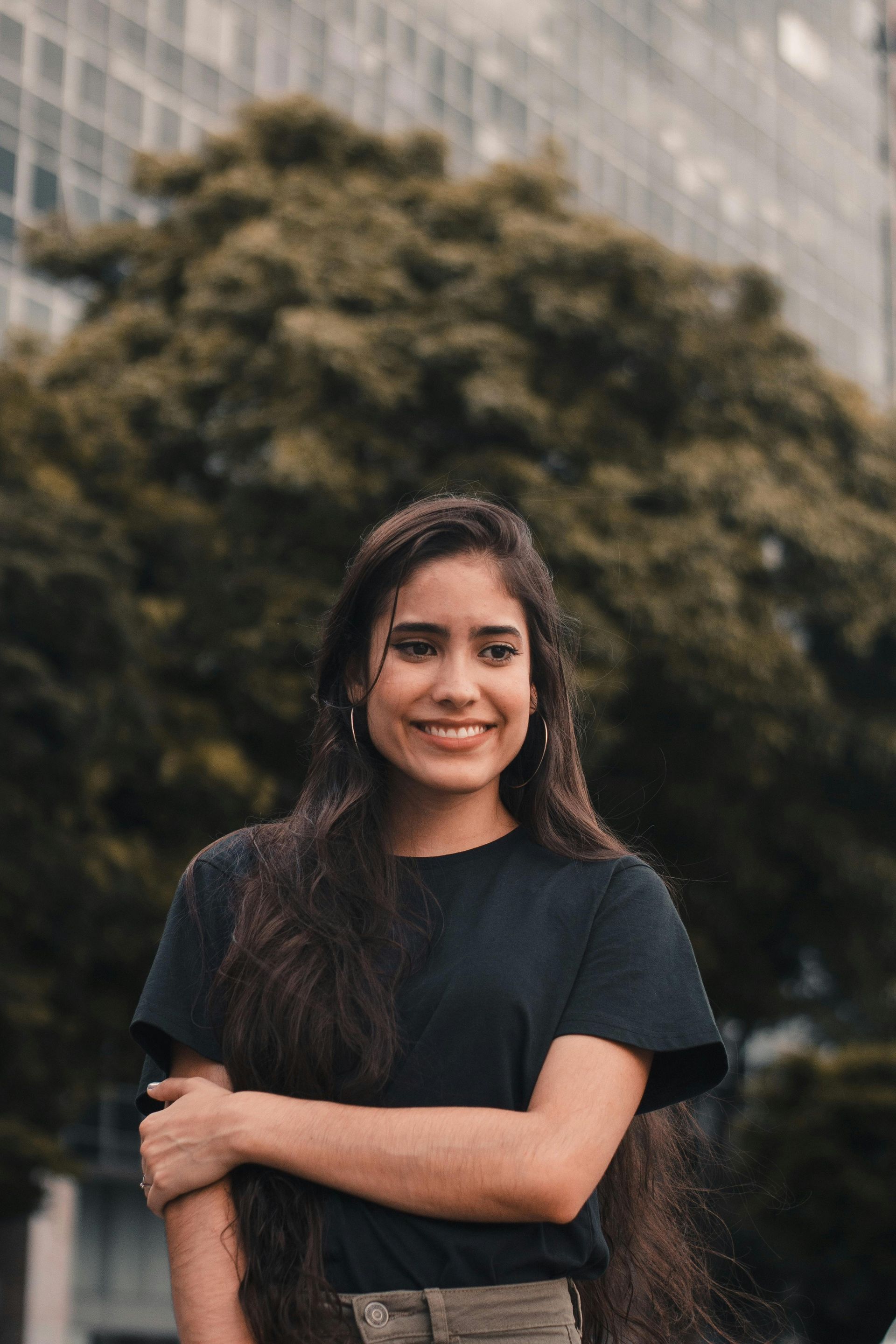 Woman in black shirt standing outdoors, smiling with arms crossed in front of trees and a building.