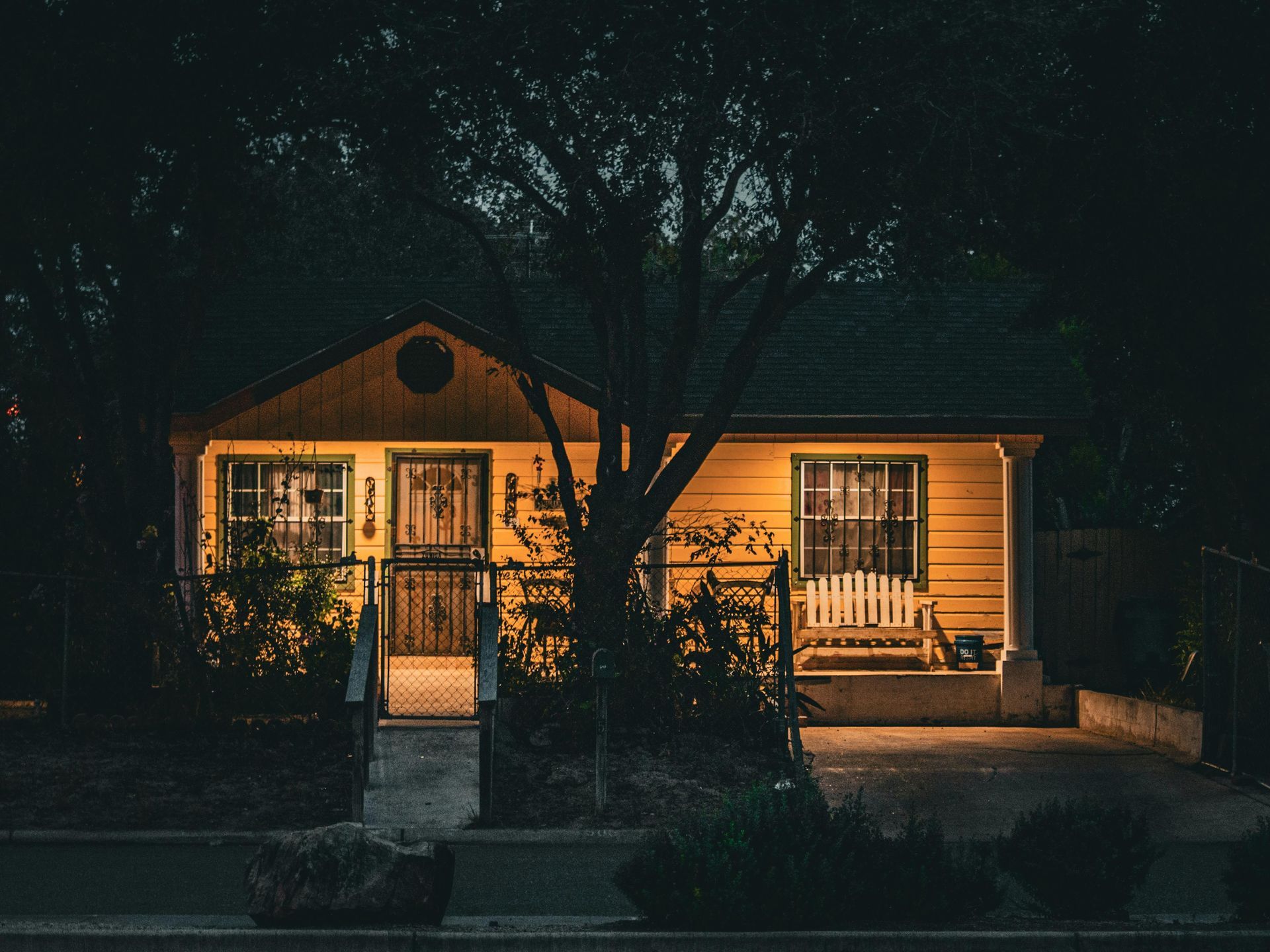 Lit front porch of a small house at night, with a tree in front and warm windows glowing.