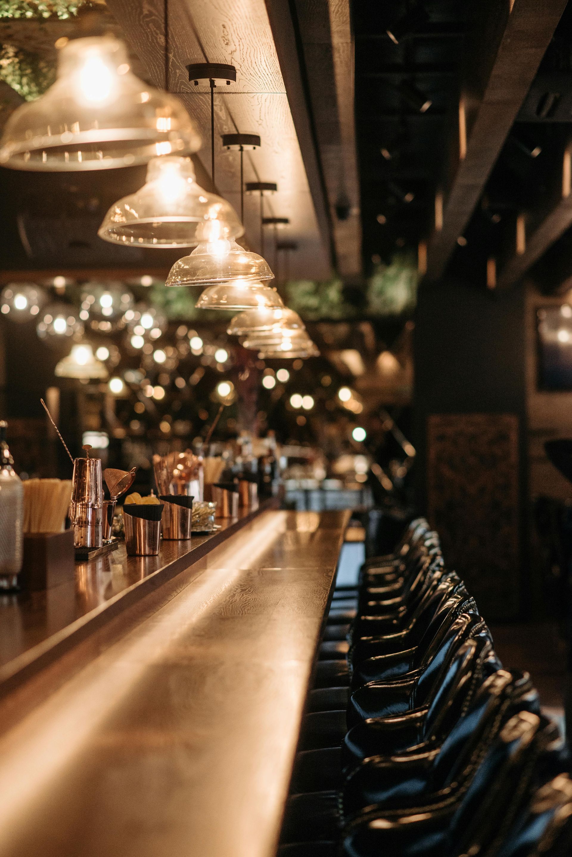 Dimly lit bar interior with hanging pendant lights, a long wooden counter, and empty barstools