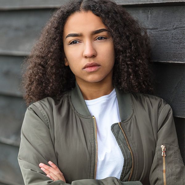 A young woman with curly hair is leaning against a wooden wall with her arms crossed.
