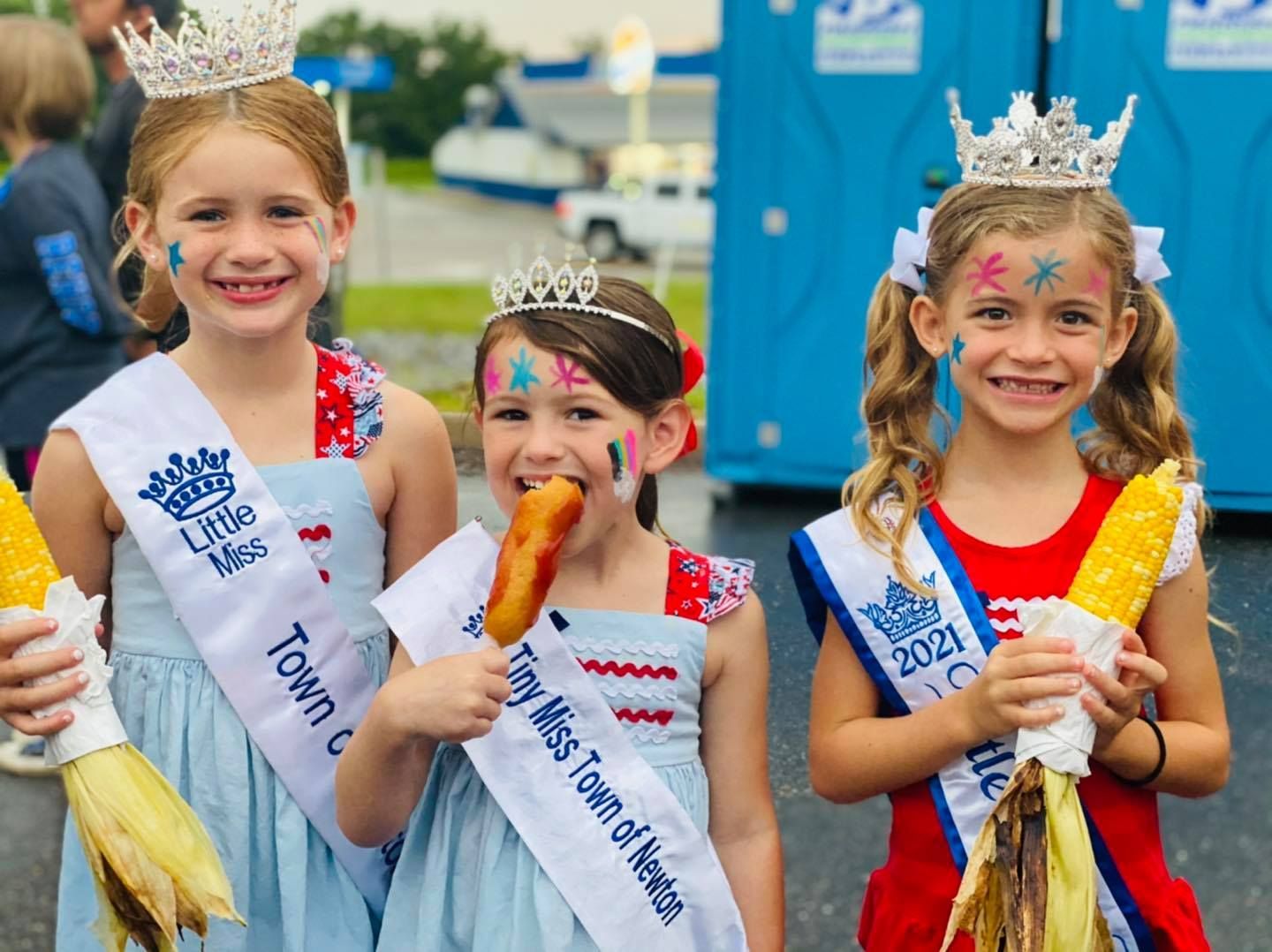 Three little girls wearing sashes and crowns are holding corn on the cob.