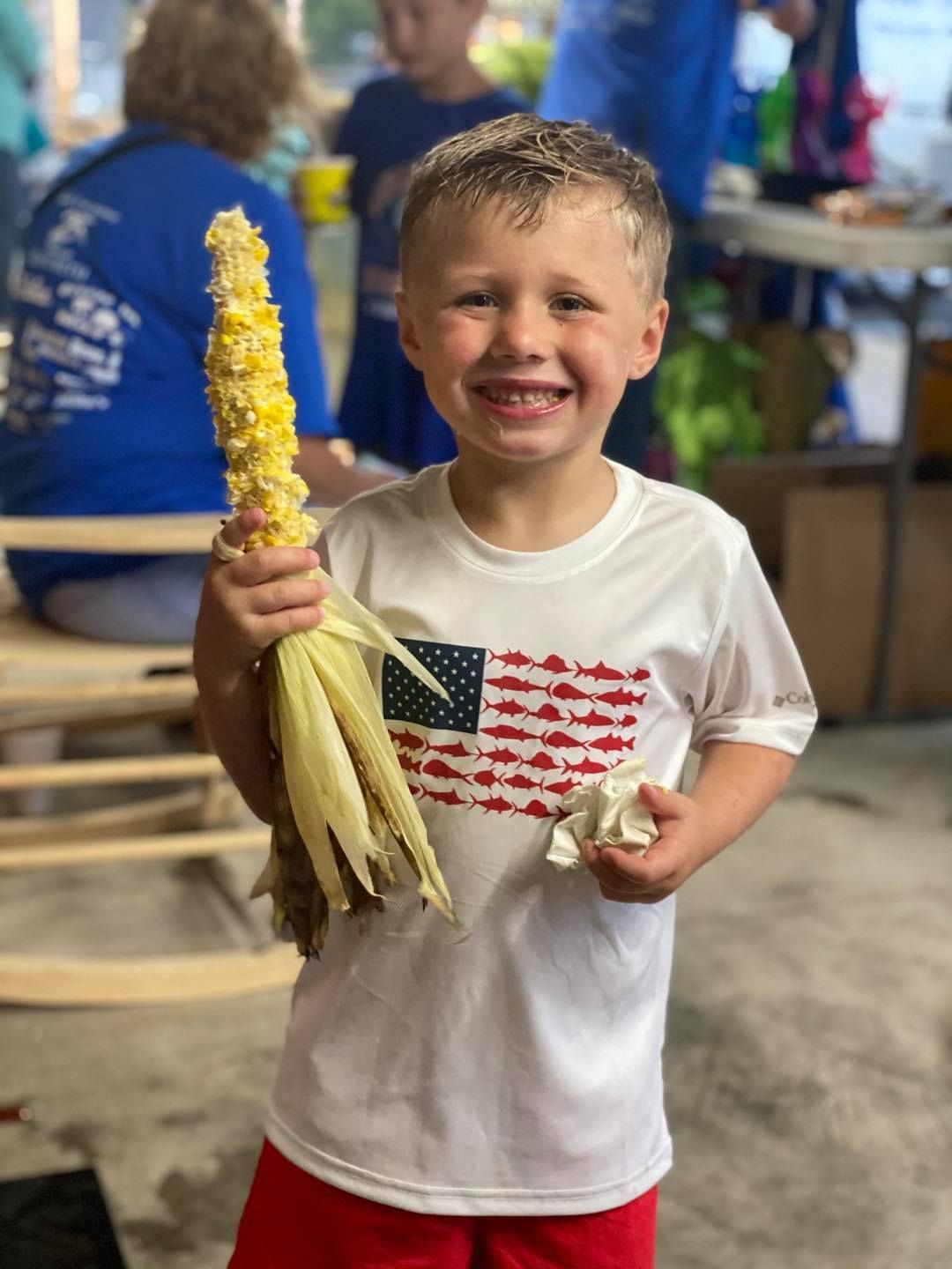 A young boy in a white shirt is holding a corn on the cob.