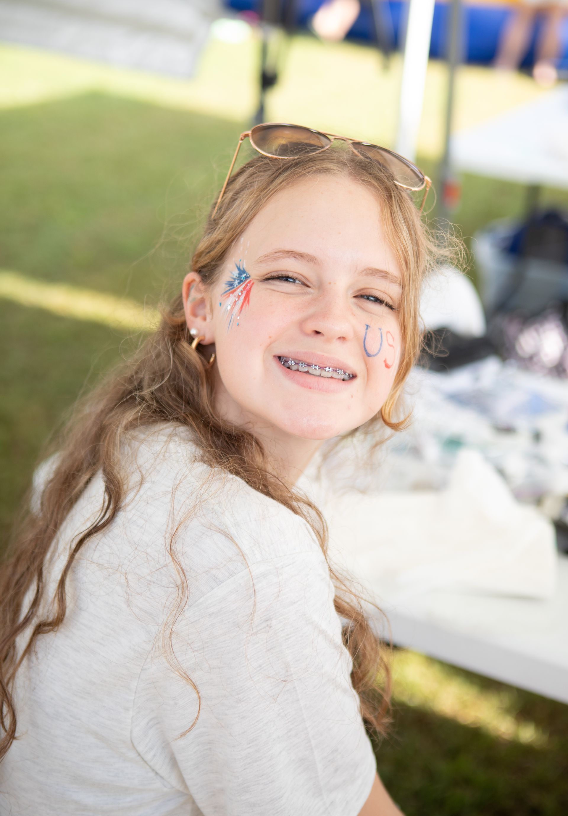 Girl with braces and face paint smiles, outdoors. Sunglasses on head, light-colored shirt.