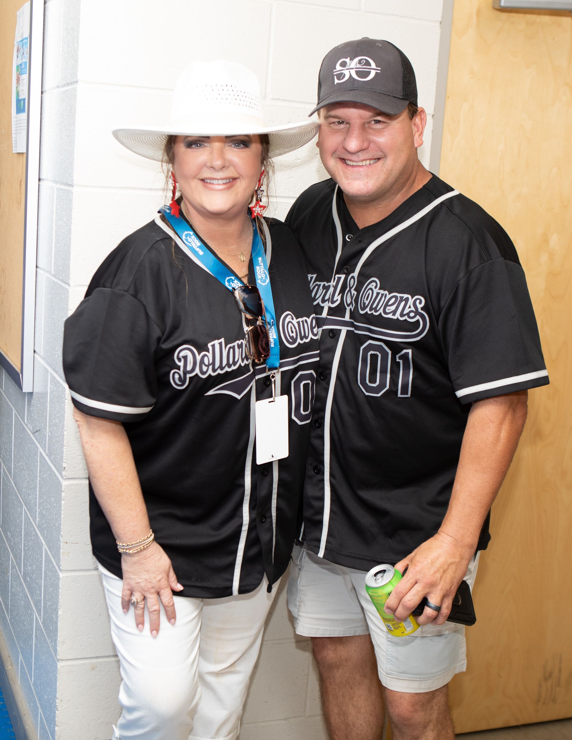 Couple wearing black baseball jerseys posing indoors. Woman wears a white hat. Man holds a can.