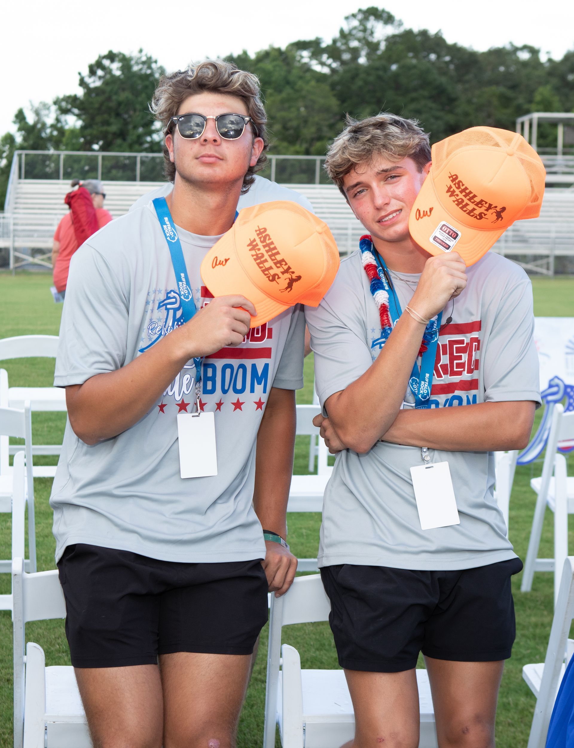 Two young men hold orange hats. They wear gray shirts, black shorts, and lanyards.  Standing on a lawn with chairs and a stadium.