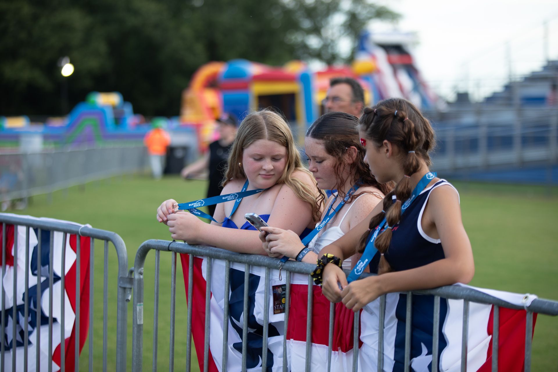 Three girls wearing blue ribbons look at a phone over a metal barrier decorated with red, white, and blue banners.