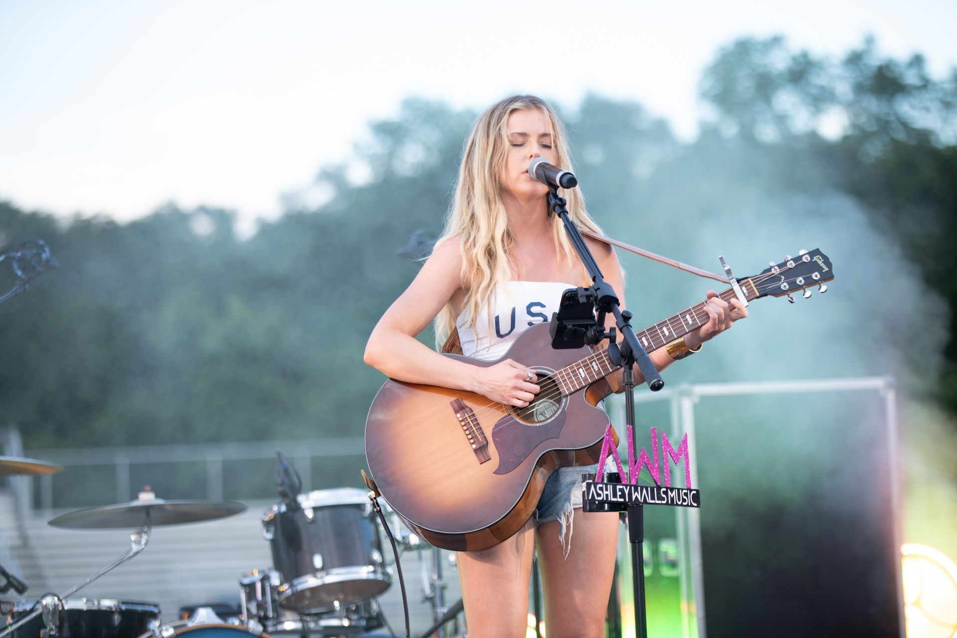 Blonde woman playing acoustic guitar and singing on stage; drums and fog in background.