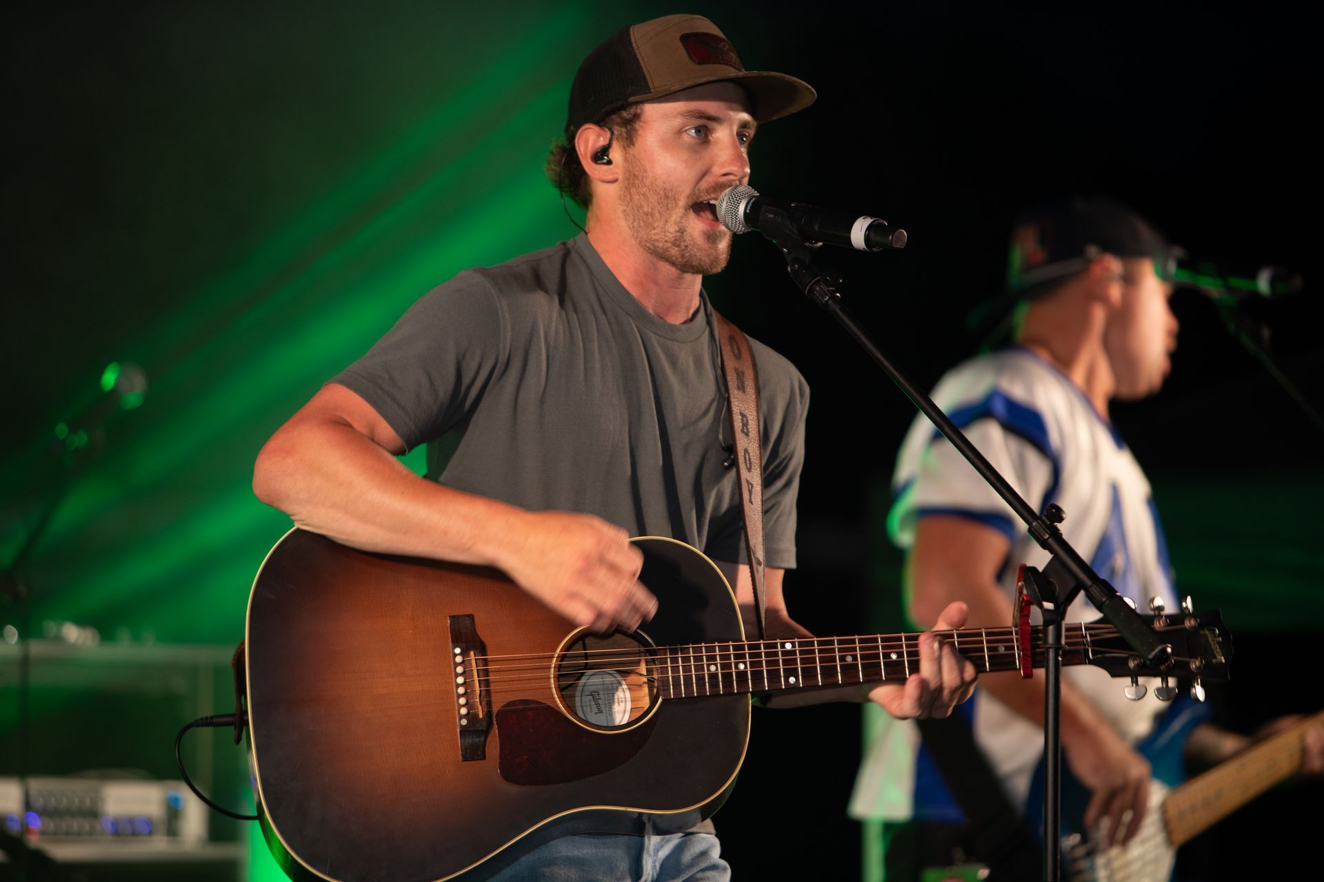 Man in baseball cap plays acoustic guitar, singing into a microphone onstage under green stage lights.