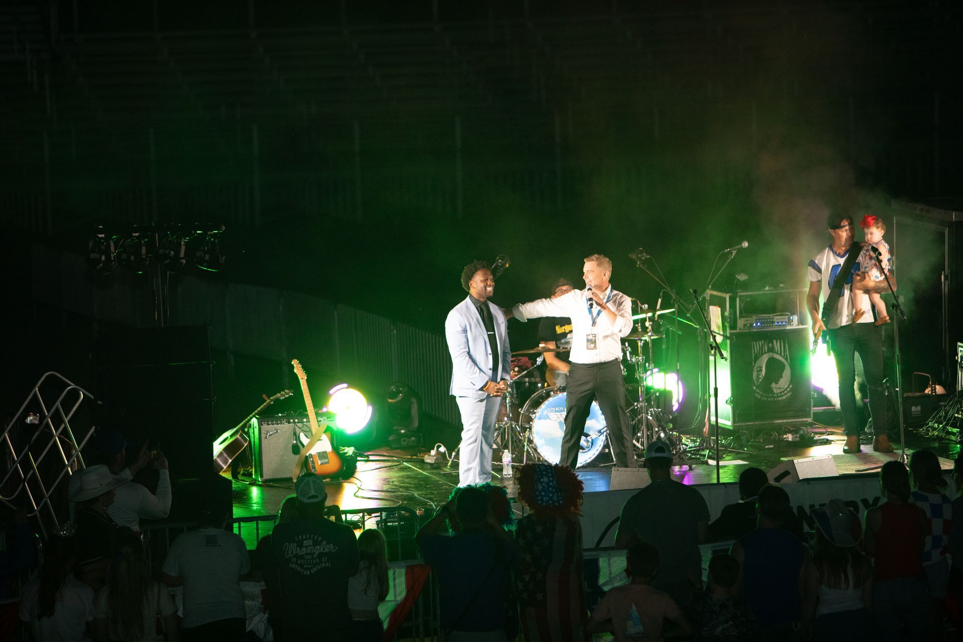 Band performs on stage, bright green lights. Singer in white jacket gestures toward another person in a suit. Audience watches.