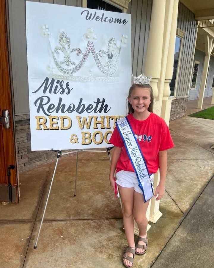 A young girl is standing in front of a sign that says miss rehobeth red white and blue.