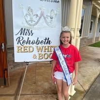 A young girl is standing in front of a sign that says `` miss rehobeth red white and blue ''.
