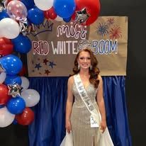 A woman in a dress and sash is standing in front of balloons.