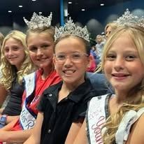 A group of young girls wearing crowns and sashes are sitting next to each other.