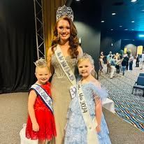A woman is standing next to two little girls wearing sashes and crowns.