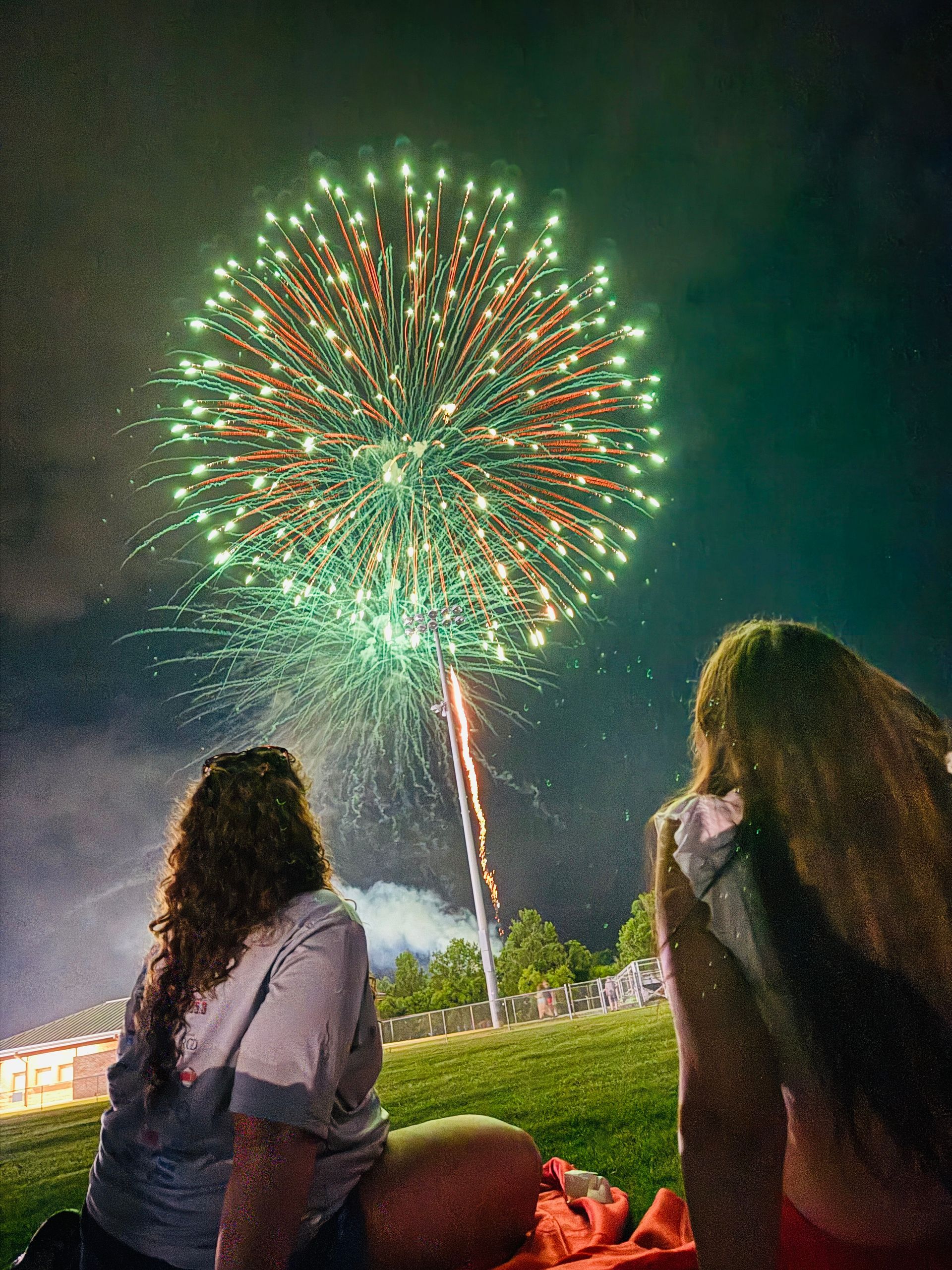 Two people watch a green firework burst over a grassy area at night.