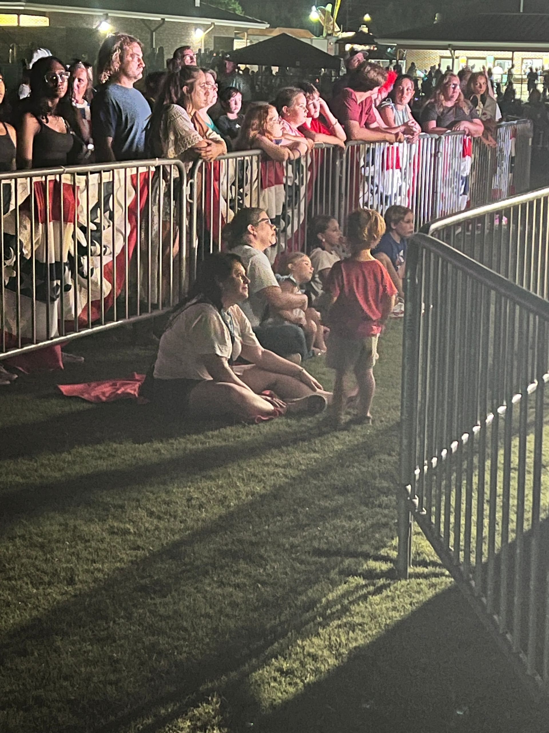 People watching an event behind a metal barrier, some sitting on artificial turf.