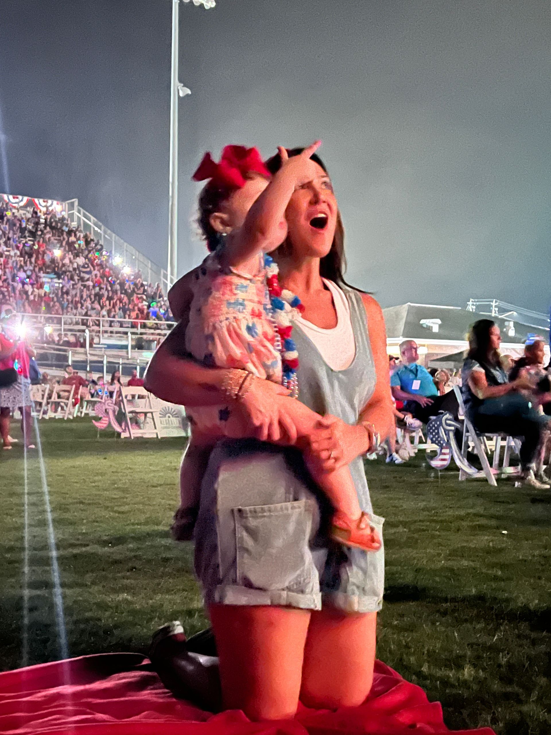 Woman holding a child, both looking up at fireworks in a stadium. Child points; both have excited expressions.