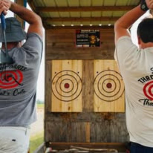 Two men are throwing axes in front of a wooden target.