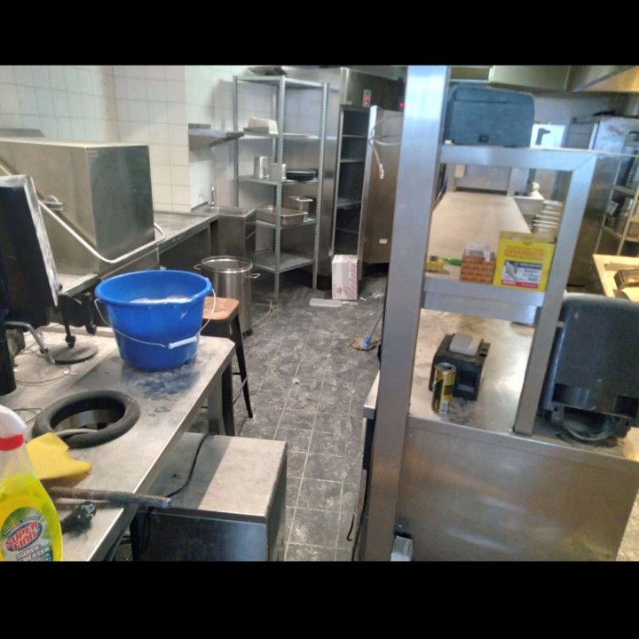A cluttered industrial kitchen with stainless steel shelving, countertops, and equipment. Floor is dirty.
