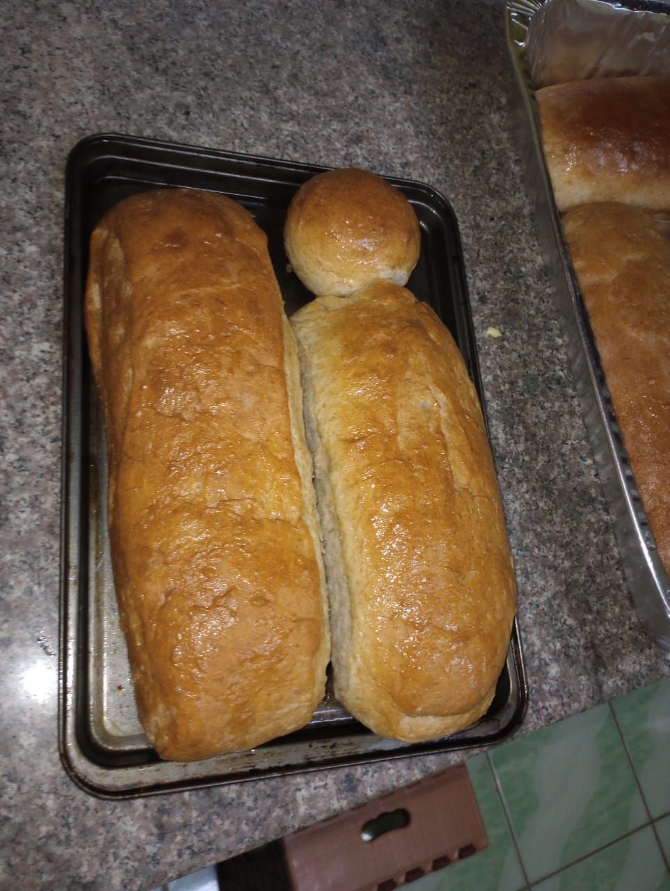 Baked loaves of bread and a roll on a baking sheet, with more loaves in a separate pan.
