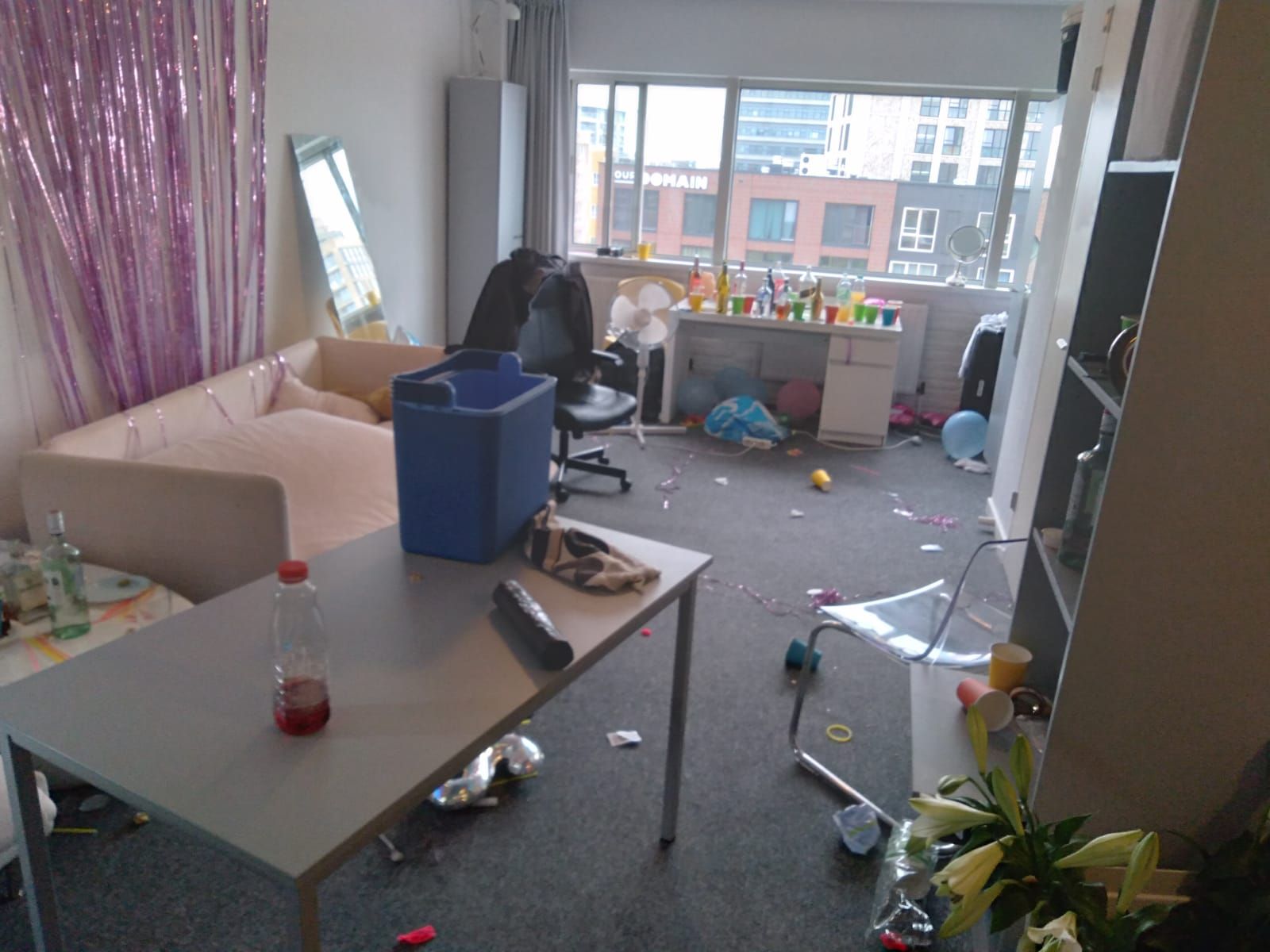 Messy living room after a party with debris scattered, table in foreground, window in background.