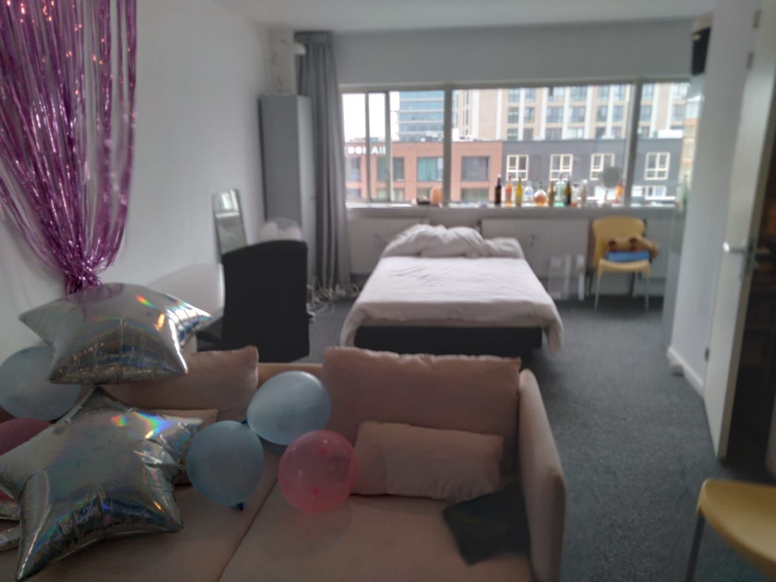 A decorated room with balloons, a bed, a couch, and a window overlooking a cityscape.
