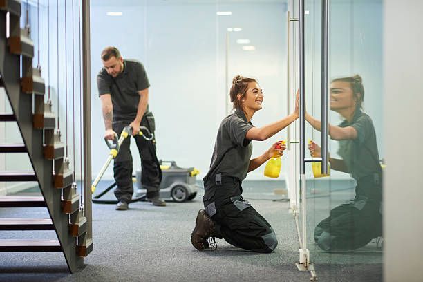 Two cleaners work in an office. One vacuums, the other cleans a glass door, smiling.