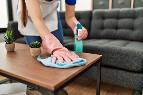 Person wearing pink gloves wiping a wooden coffee table with a blue cloth, spraying it with cleaner in a living room.