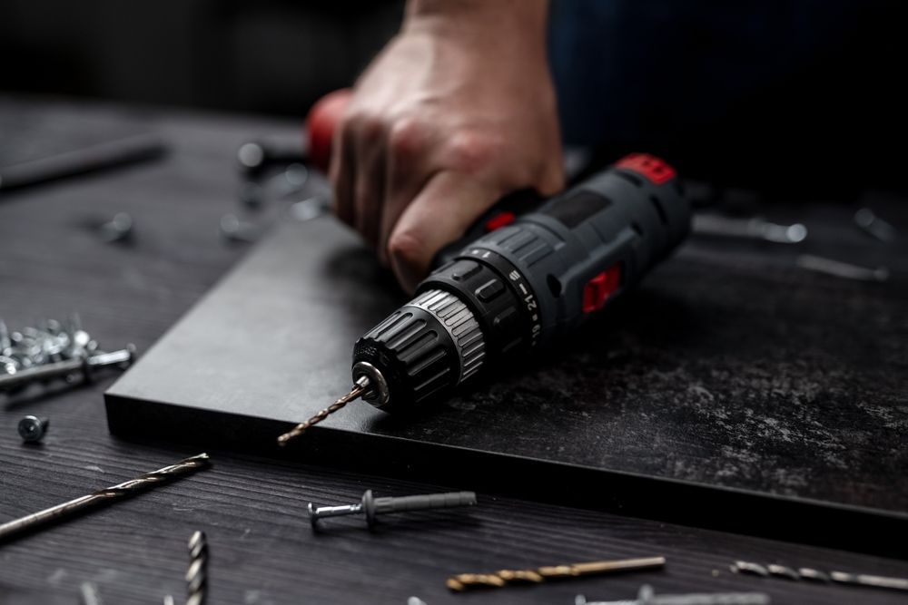 Hand holding a power drill, drilling into a dark surface; various drill bits and screws are scattered around.