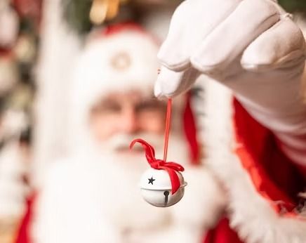 Santa Claus in a red suit and white gloves holds up a white jingle bell ornament tied with a red ribbon.