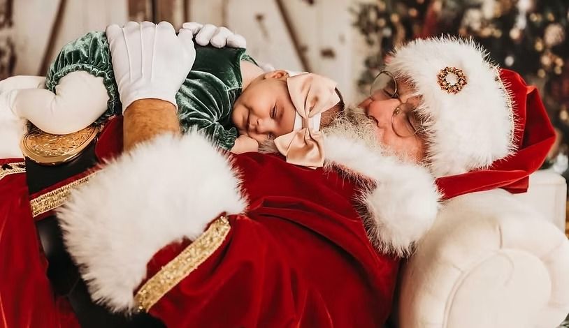 Santa Claus, in a red suit and white fur trim, gently holds a baby wearing a green velvet outfit and tan bow headband.