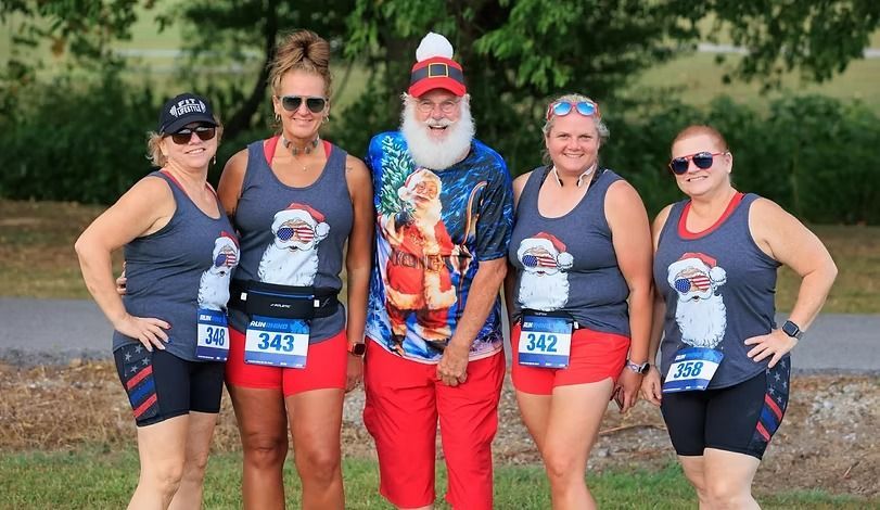 A group of five people in patriotic Santa-themed athletic wear pose for a photo in a park setting.