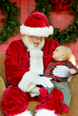 Santa Claus in a red suit sitting in an armchair holding a baby who is wearing a plaid shirt and looking at him.