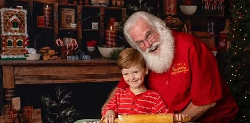 Santa and a young child smiling while rolling out dough in a decorated kitchen setting.