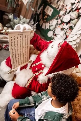 Santa Claus in a red suit reads a long scroll to a child in a plaid shirt with a Christmas tree in the background.