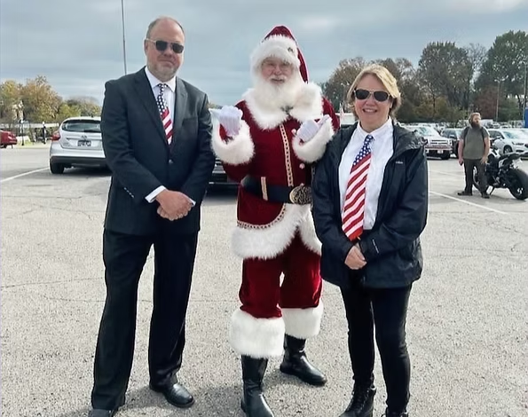 A man in a suit, a person dressed as Santa Claus, and a person with an American flag tie pose together in a parking lot.