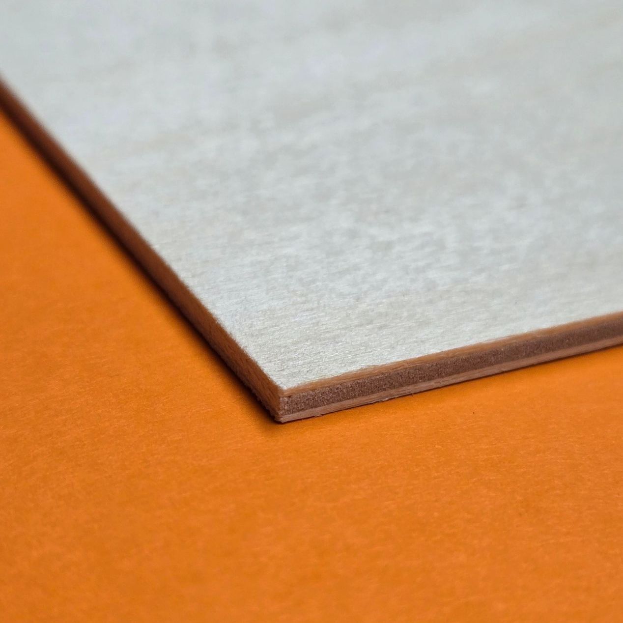Light birch plywood sheet on orange background, showcasing fine grain layers and clean-cut corner profile.