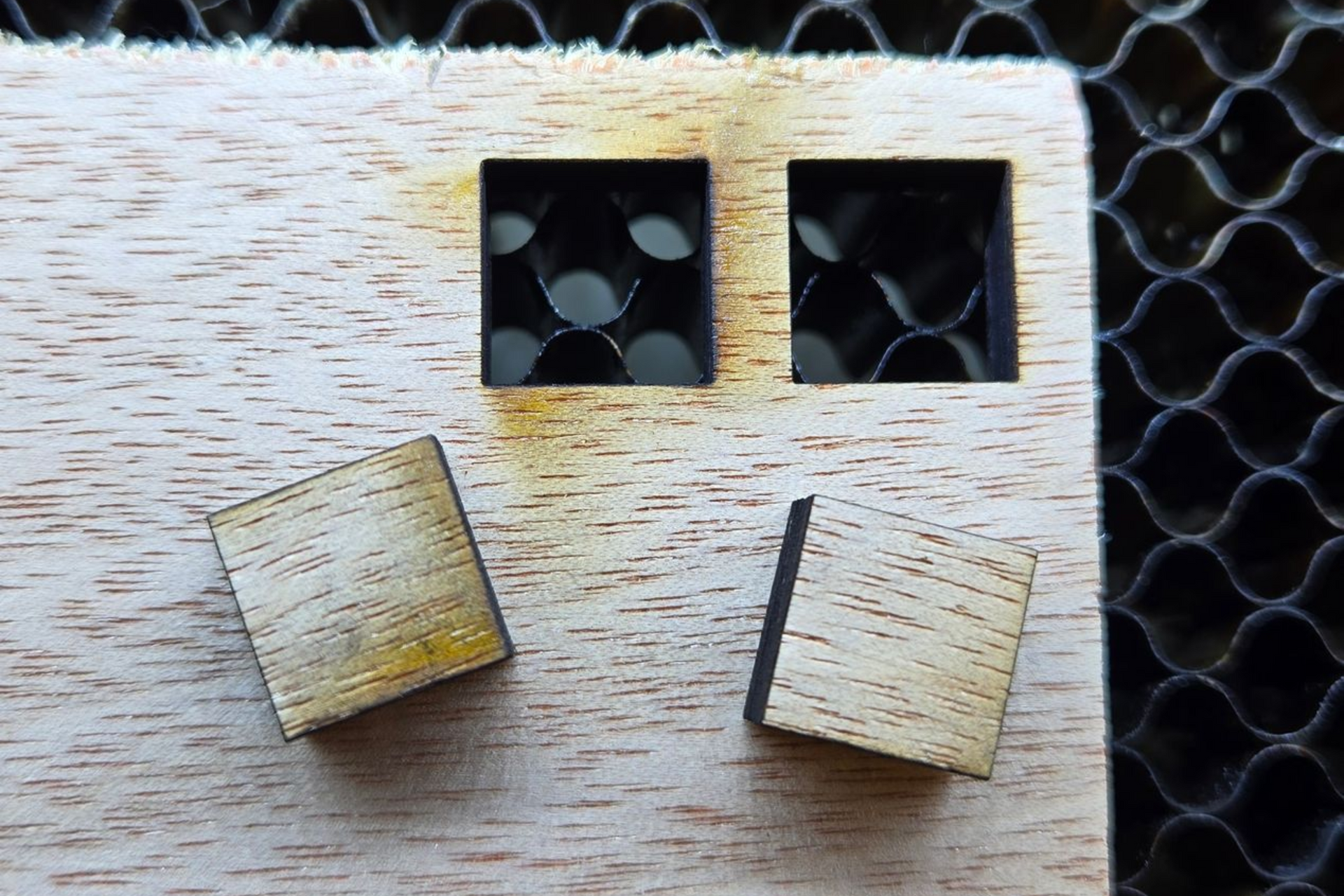 Close-up of laser-cut wood squares with and without proper air assist showing edge quality on a honeycomb laser bed.