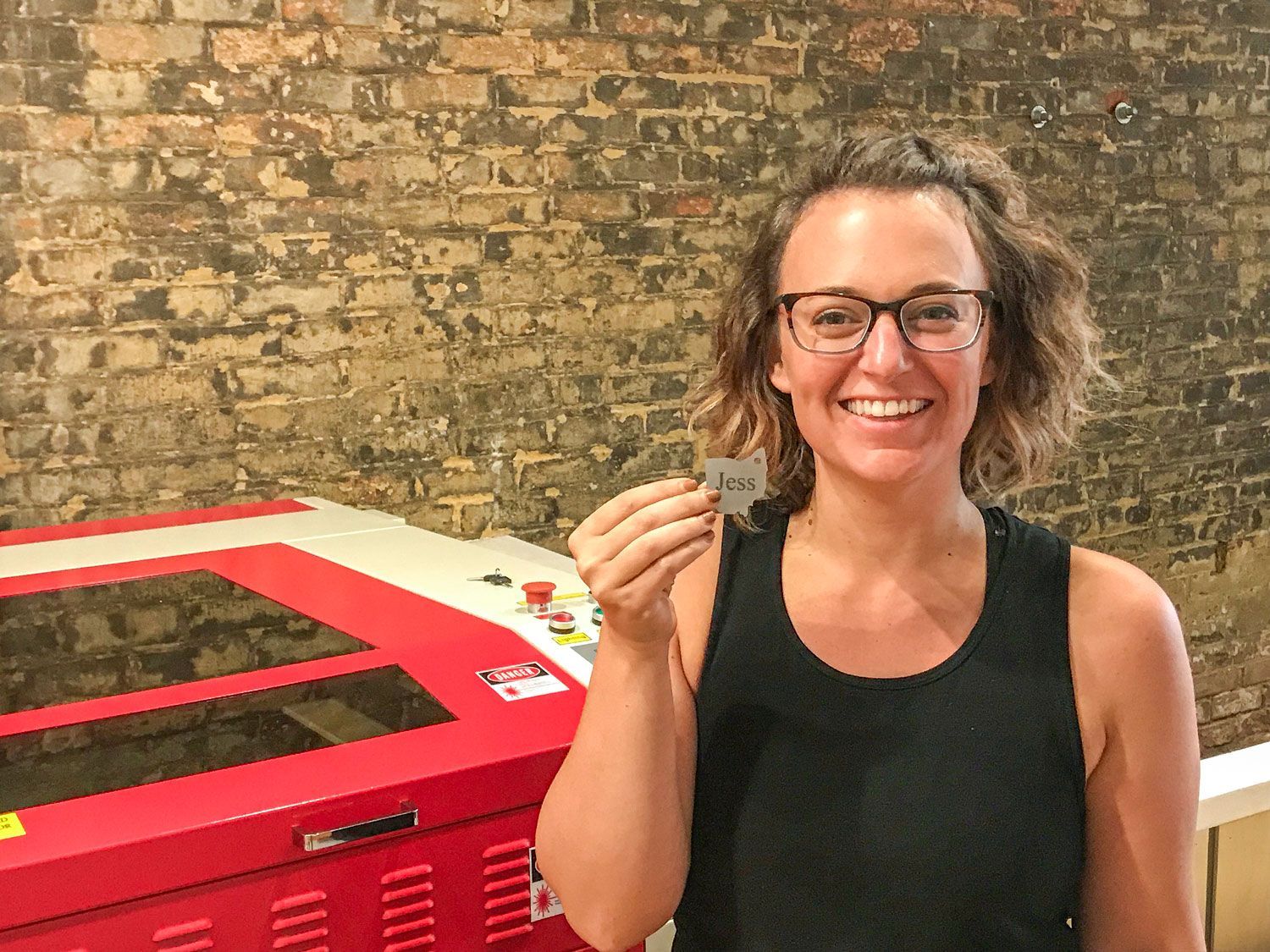 Smiling woman in glasses holds up her first laser-cut project with her name “Jess” engraved, standing beside a red laser machine.