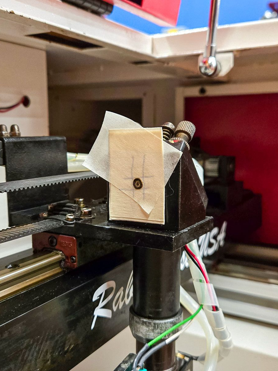 Close-up of a laser mirror mount with masking tape showing a centered burn mark for beam alignment inside a laser machine.